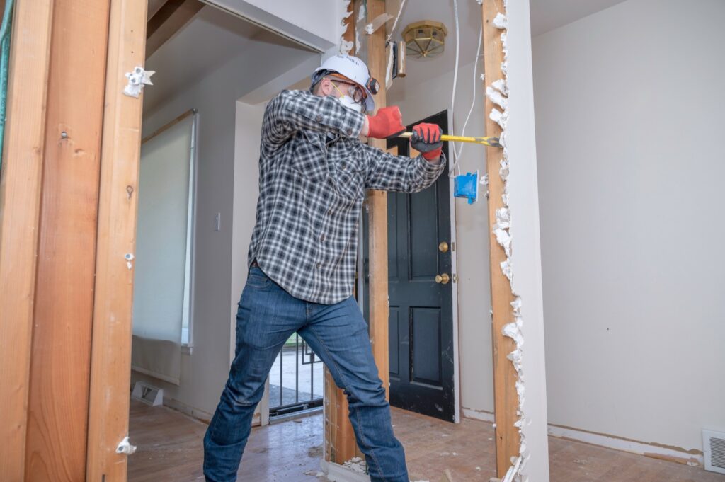 Worker in a dust mask prying drywall off a wall to expose the wood stud framing behind it
